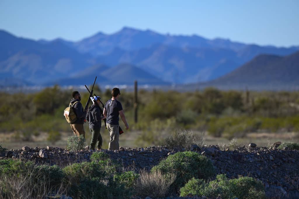 Three people walking through a desert landscape with shotguns
